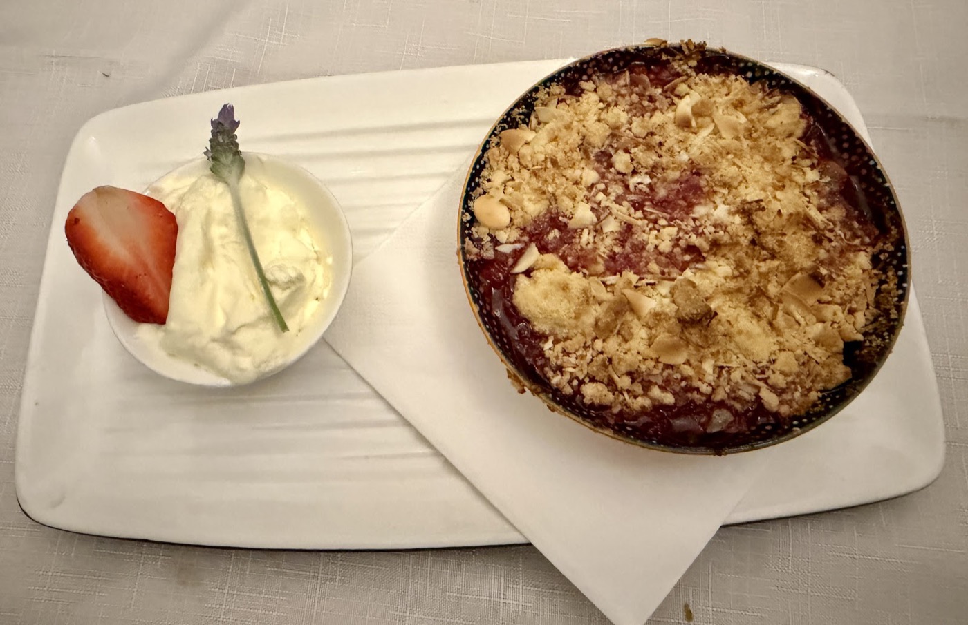 A baked crumble in a small round dish beside a pot of cream topped with a strawberry half and a sprig of lavender, on a white plate