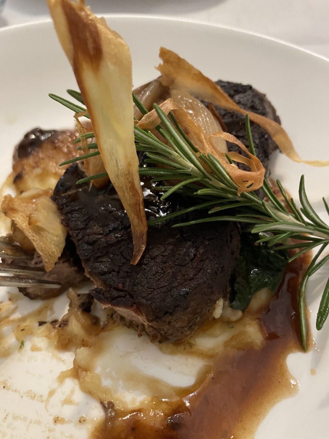A seared beef fillet on creamy potato with a rosemary sprig, crisp strips on top and a dark sauce