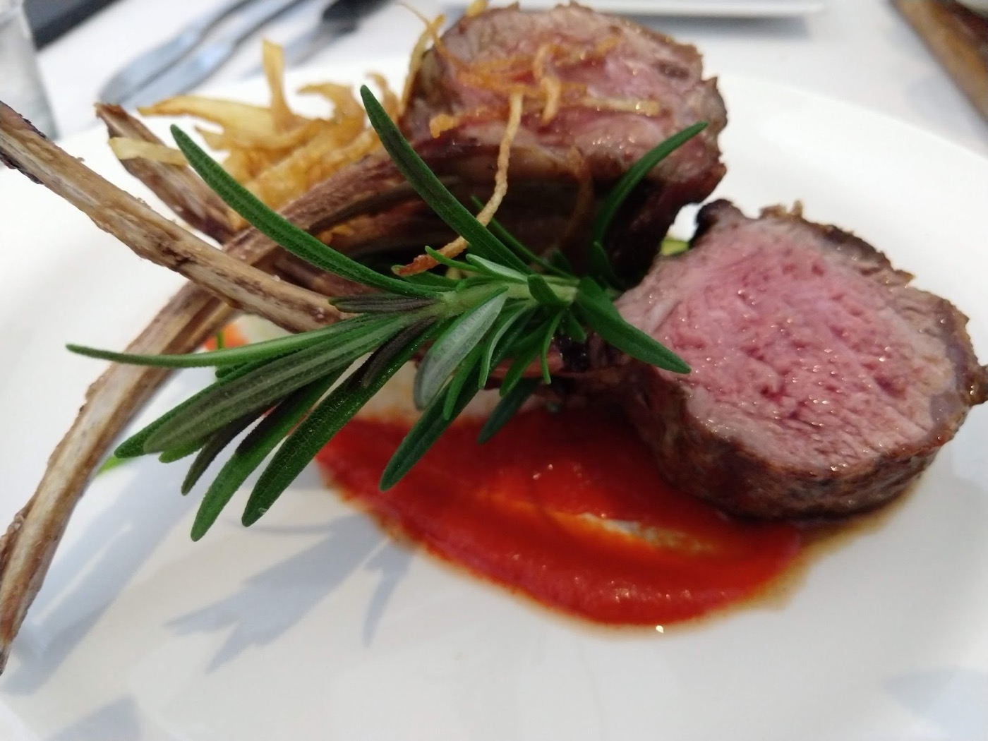 Two pieces of pink lamb on a streak of red sauce with a rosemary sprig and fine fried potato strands