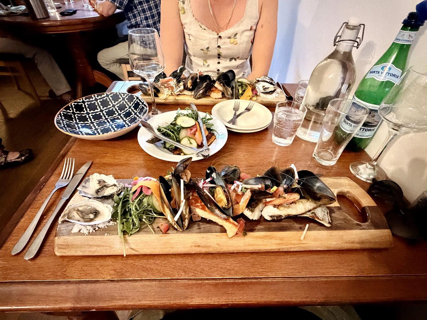 A wooden board of oysters, mussels and other seafood on a timber table, with a side salad and water bottles