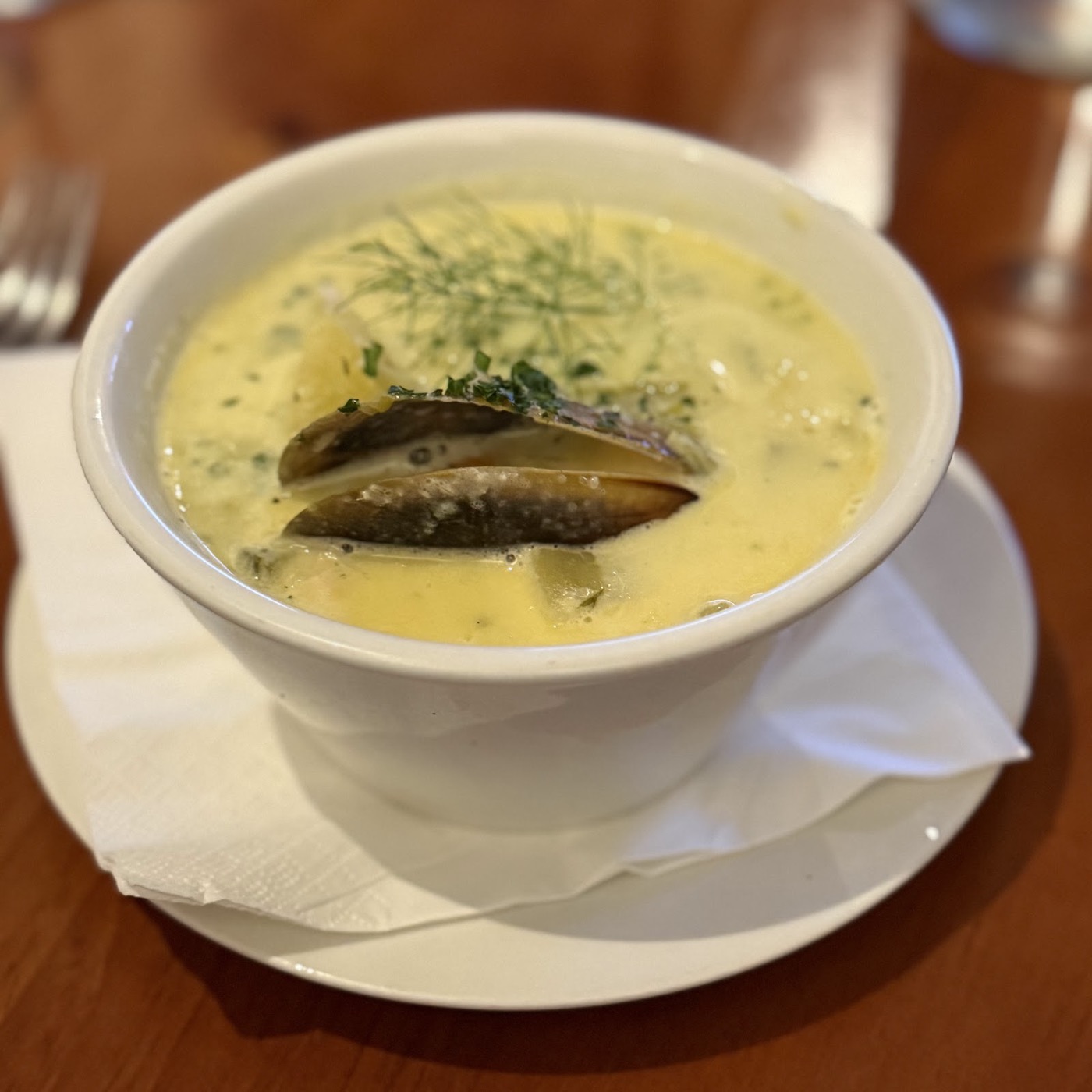 A white cup of creamy yellow chowder with a mussel shell and fennel on top, on a saucer with a folded napkin