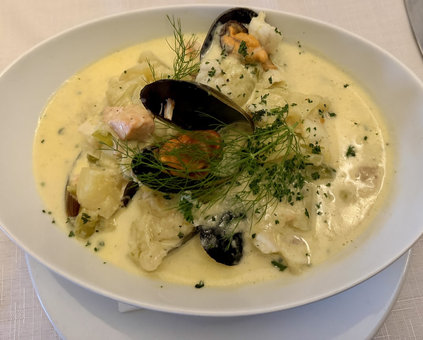 A wide white bowl of creamy seafood chowder with mussels, fennel fronds and fresh herbs